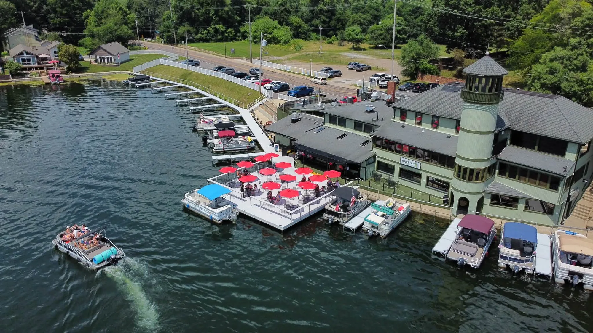 Aerial view of Harbor Lighthouse Grille: the green lighthouse-tower building on the shore of Portage Lakes, with red patio umbrellas and boats tied up at the docks