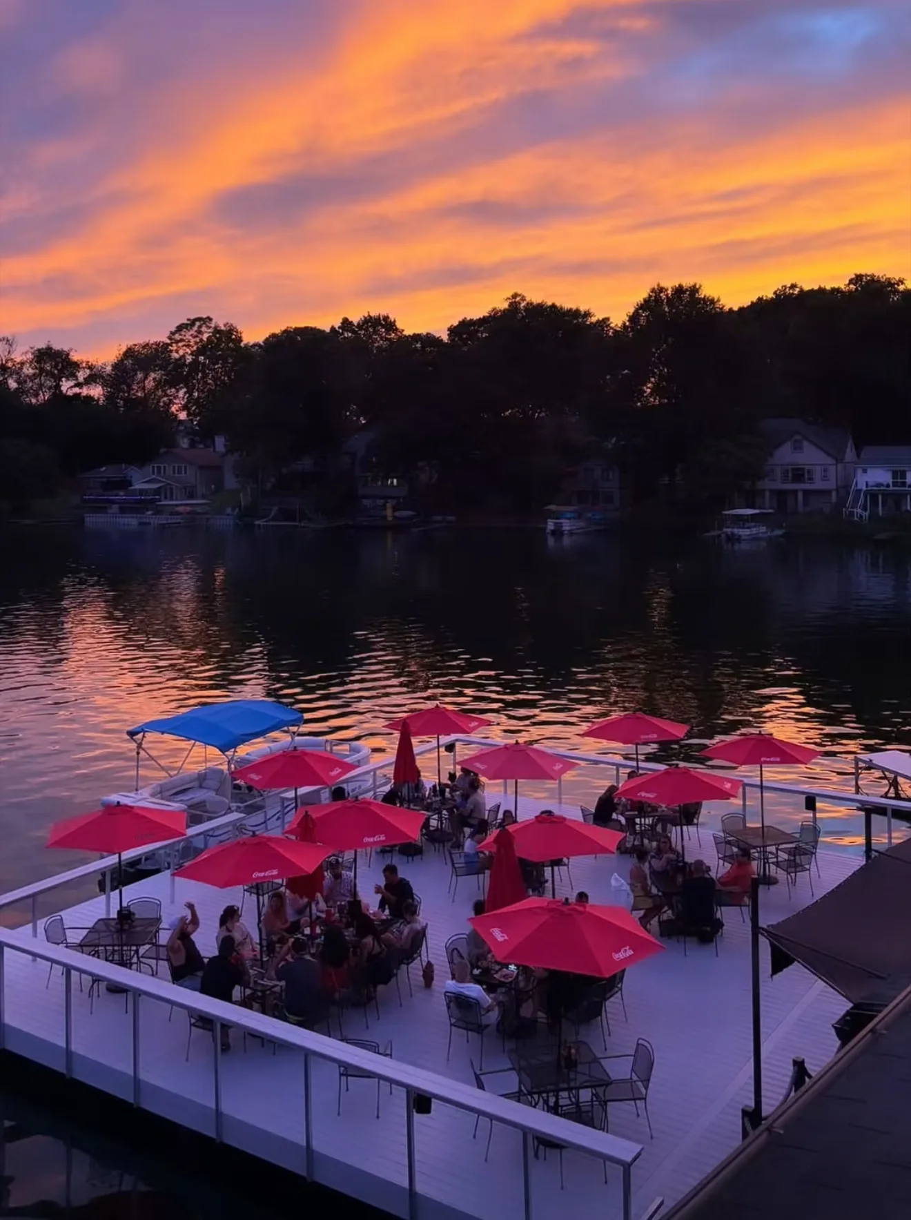 A full deck of guests under red umbrellas on the floating patio at Harbor Lighthouse Grille, an orange sunset over Portage Lakes behind them